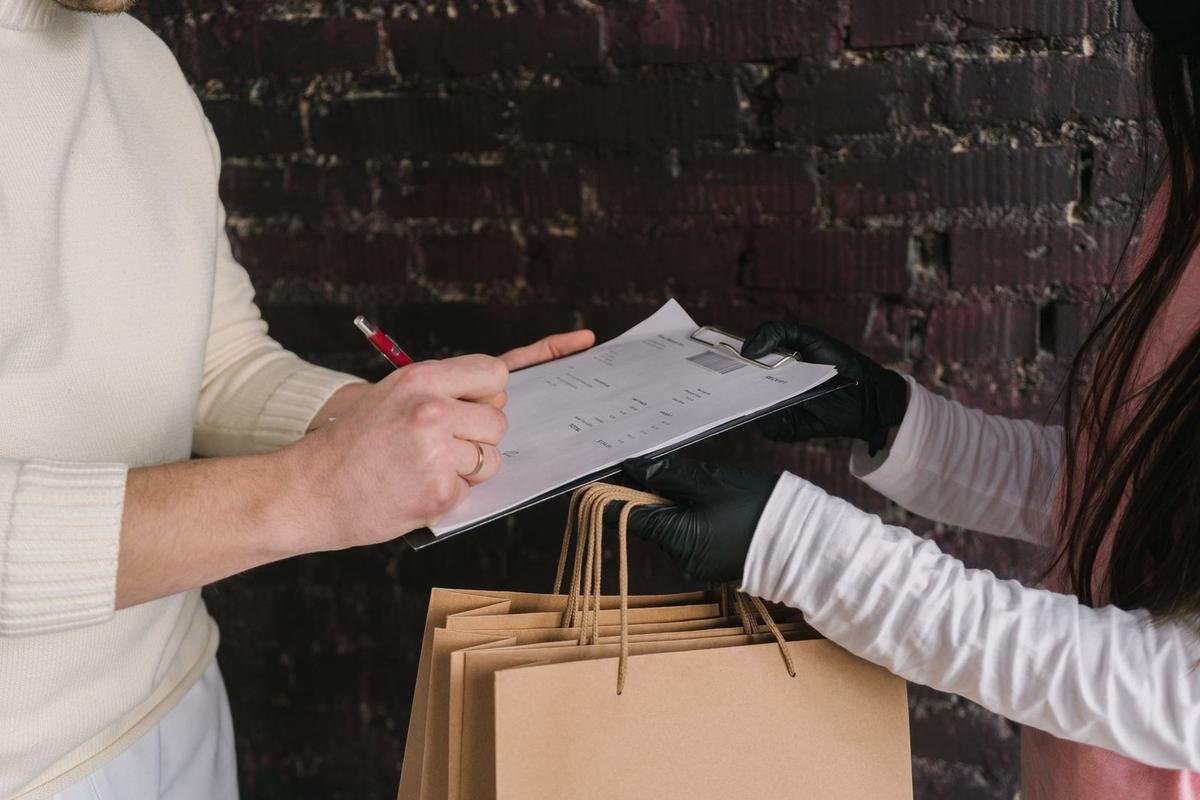 Deux individus signent un document, l'un avec des sacs en papier, contre un fond de mur en briques